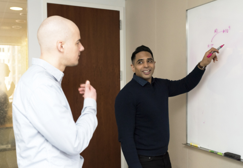 One man explains something on a whiteboard to another man at his company.